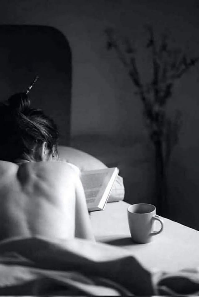 A woman from behind sitting in bed with a book and a mug in soft grey light